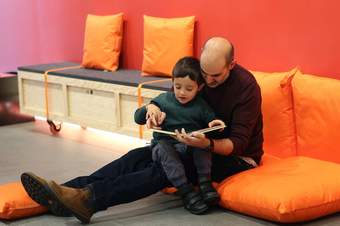 A photograph of a father and son sat on cushions on the floor whilst reading a book