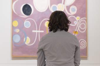 A person with shoulder length dark hair sitting looking at a lilac abstract painting