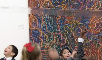 Children look up at a drawing made on a large sheet of transparent plastic which is hung above their heads. An artwork with coloured squiggly lines is in the background.