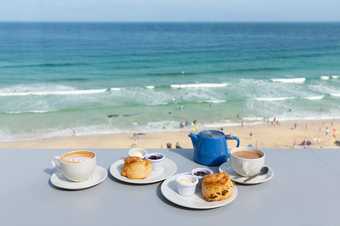 a cup and saucer, two plates with scones cream and jam and a blue teapot are laid on a greay surface. Behind are views of the sea and sky. 