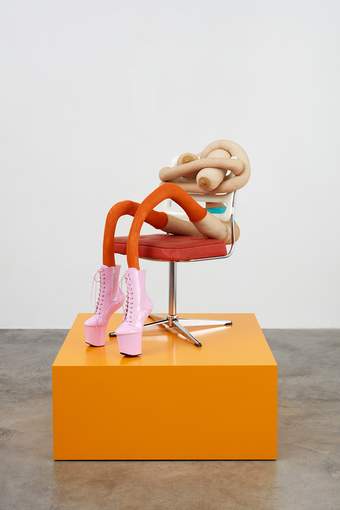 Seated Sarah Lucas sculpture wearing pink platform boots on a square orange plinth