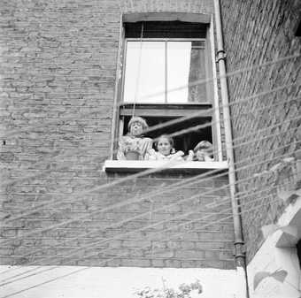 Girls look down from a first floor window of a brick house, with washing lines in front of them